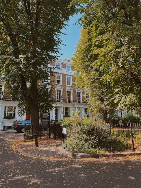 A long, straight outdoor walkway lined with tall trees on both sides, their branches forming a canopy overhead. The ground is covered with fallen yellow and brown leaves, creating a layered carpet. Along the pathway, there are vintage-style black lampposts and small metal bollards to manage pedestrian and vehicle movement. In the background, the pathway extends towards a bright, sunlit horizon, with a few distant figures visible at the far end. On the right side, there are several low-rise buildings and parked cars, indicating a residential or urban environment near Holland Park gardens. The scene is illuminated by natural daylight, casting soft shadows and highlighting the vibrant autumn foliage. This peaceful setting could be part of a park or public space where house removals and furniture transport could be coordinated, with Removal Company Holland Park potentially managing the logistics of home relocation within such tree-lined environments.