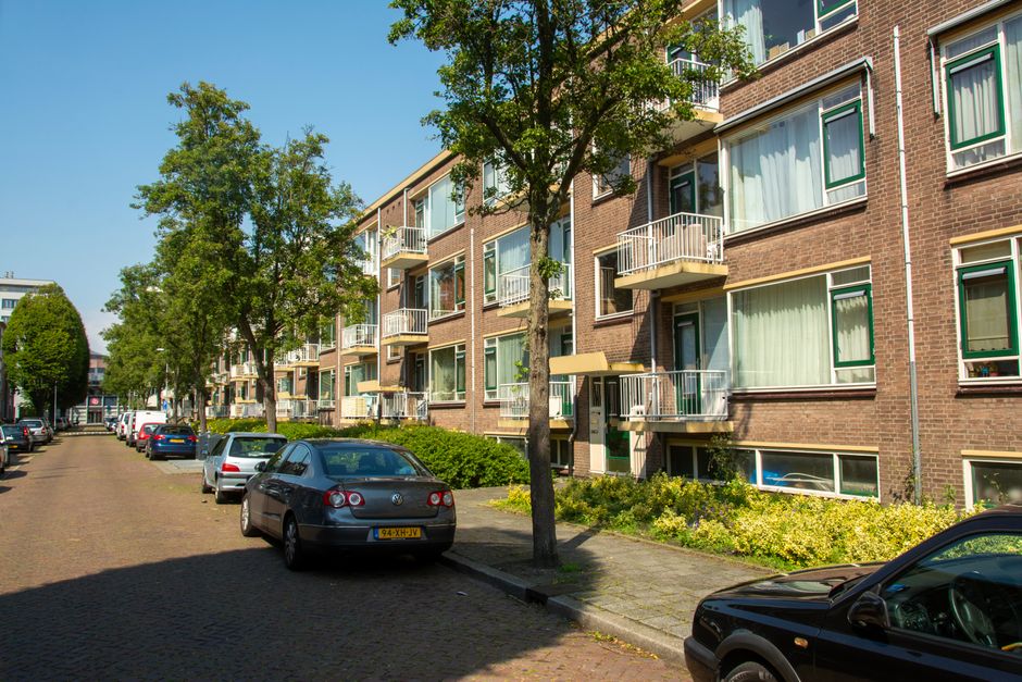 A narrow residential street in Holland Park with a row of terraced houses on the left side, featuring white walls, bay windows, and small front gardens. Parked cars line both sides of the brick-paved 
