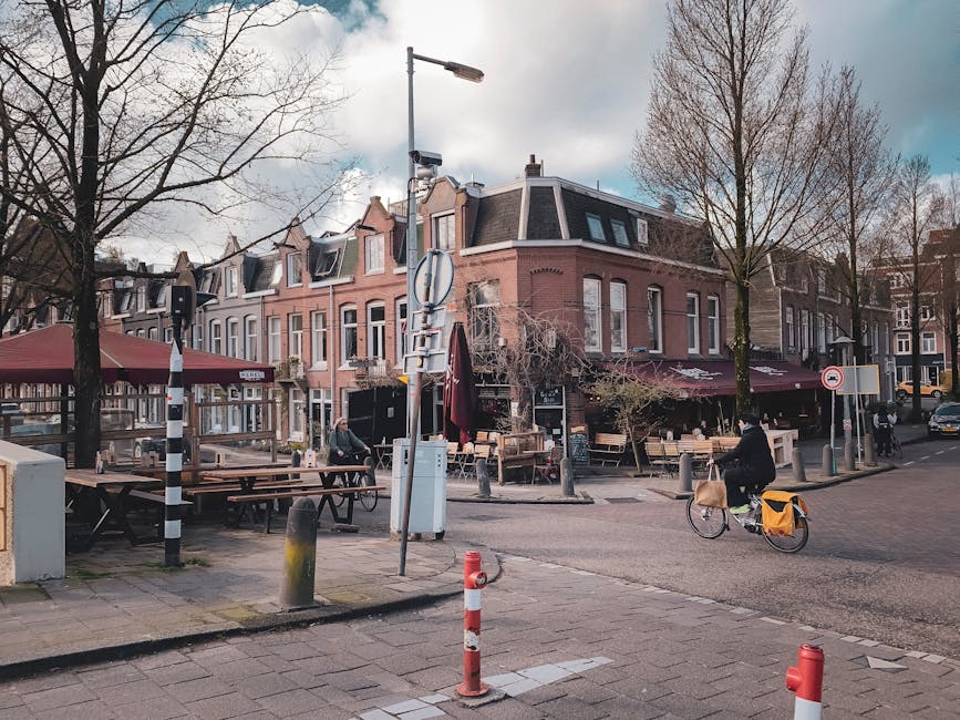 An exterior view of a three-story corner building in Holland Park during daytime, featuring brick and stucco facades, with large ground-floor windows and outdoor seating areas of a café under maroon awnings. The sidewalk in front is equipped with wooden benches, tables, and umbrellas; some are folded down. Two leafless trees are present on the sidewalk, and a black-and-white striped traffic bollard is visible near the curb. A city light pole stands close to the building, supporting traffic signs and a CCTV camera. A cyclist with a yellow pannier bag is riding past the café on a paved street, with additional parked bicycles and a parked car visible in the background. The sky is partly cloudy, and the overall scene conveys a typical urban environment suitable for house removals or relocation services by companies like Removal Company Holland Park.